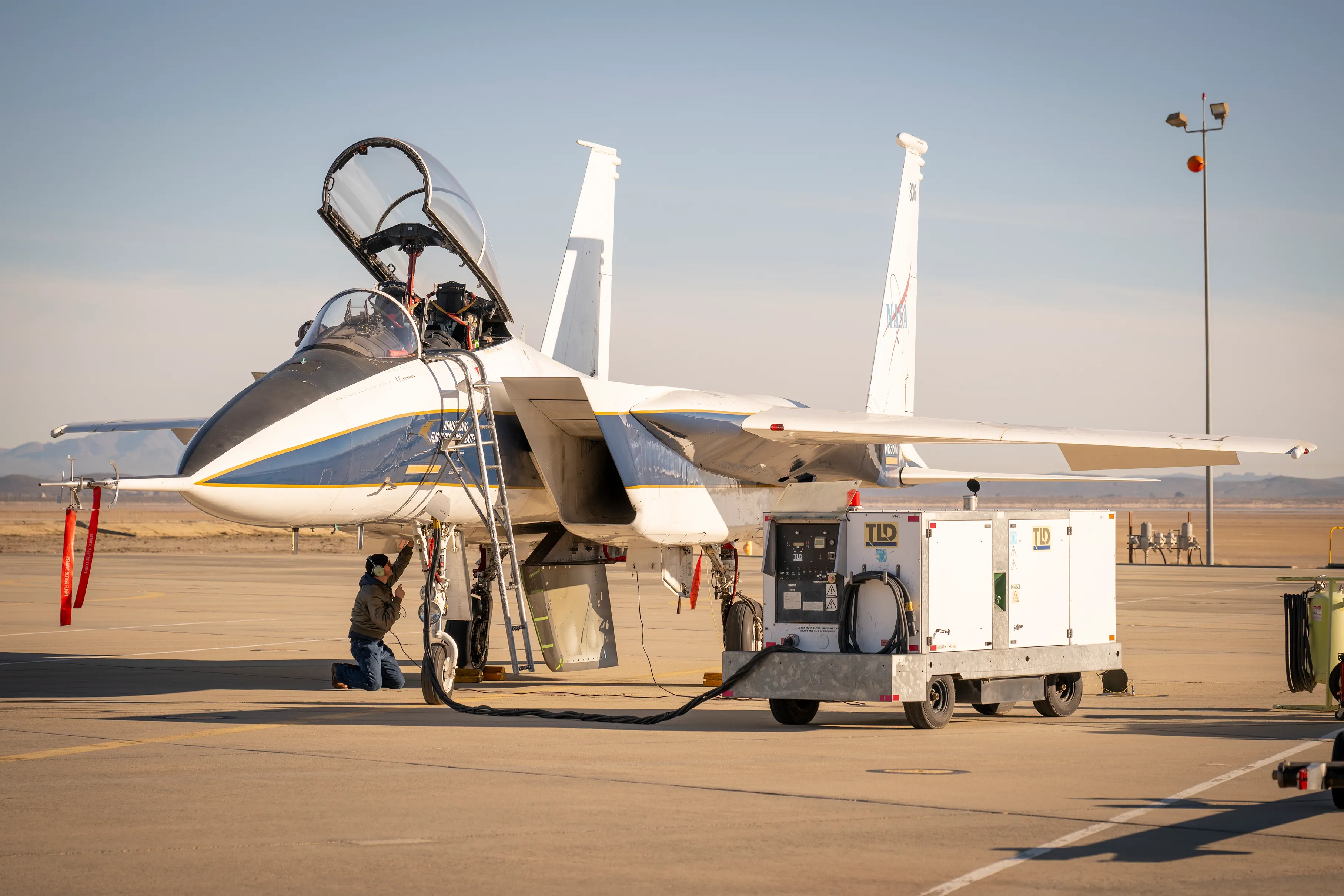 NASA'nın F-15 araştırma uçağı, NASA'nın Armstrong Uçuş Araştırma Merkezi'nde bir rampada duruyor. Temel ekipler, uçağın altında, bir deneysel test makalesi üzerinde çalışıyor, bu makale bir ventral fini andırıyor.