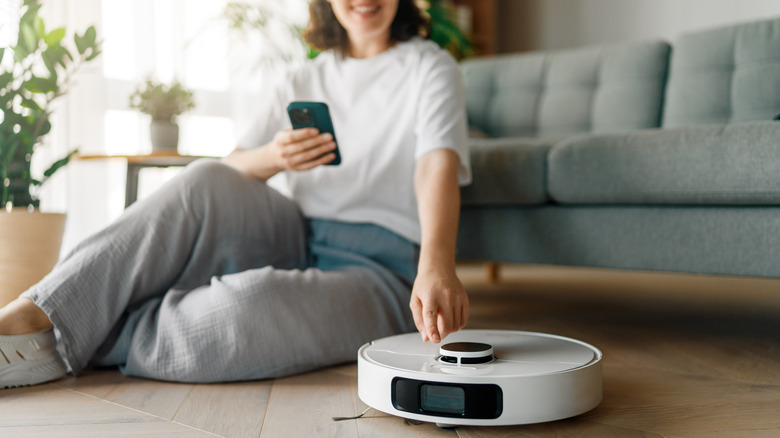 Bir kadın yerde oturmuş telefonunu tutarken gülümsüyor, önünde bir robot süpürge